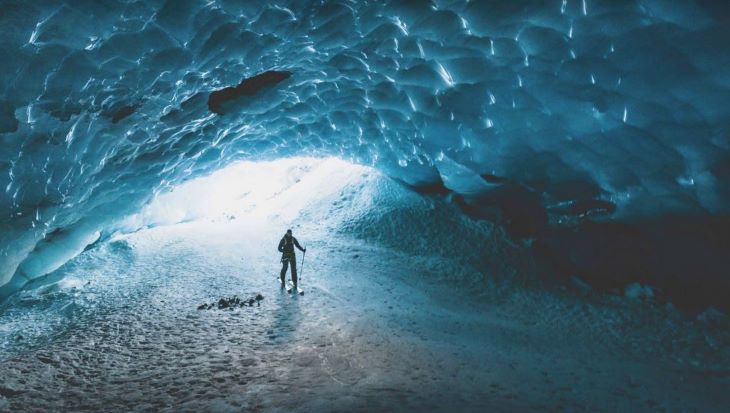 Eine Person steht in einer beeindruckenden Eishöhle, die von blauen Eiskristallen umgeben ist, während ein heller Lichtschein von der Höhlenöffnung hereinfällt.